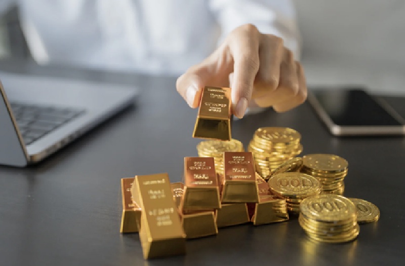 Man holding a small gold bar in his finger that is piled next to other bars and gold coins